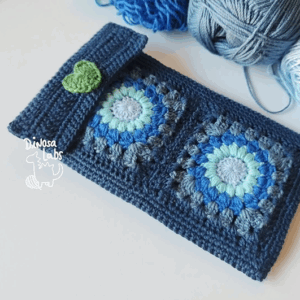 Funda para móvil hecha con crochet de colores azules y detalle de cierre en forma de corazón verde.