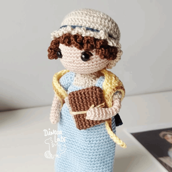 Amigurumi tejido en crochet de Jane Austen, con un vestido azul, una bufanda amarilla y un libro en la mano.