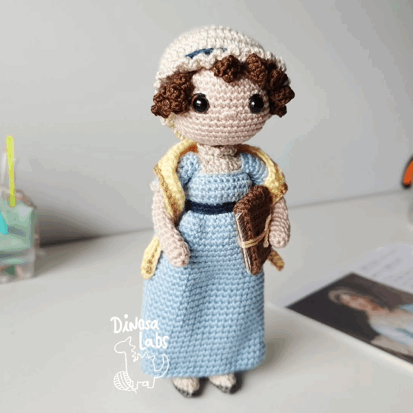 Amigurumi tejido en crochet de Jane Austen, con un vestido azul, una bufanda amarilla y un libro en la mano.