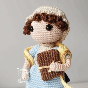 Amigurumi tejido en crochet de Jane Austen, con un vestido azul, una bufanda amarilla y un libro en la mano.