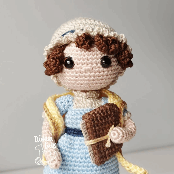 Amigurumi tejido en crochet de Jane Austen, con un vestido azul, una bufanda amarilla y un libro en la mano.