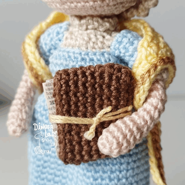 Detalle del Amigurumi tejido en crochet de Jane Austen, con un vestido azul, una bufanda amarilla y un libro en la mano.