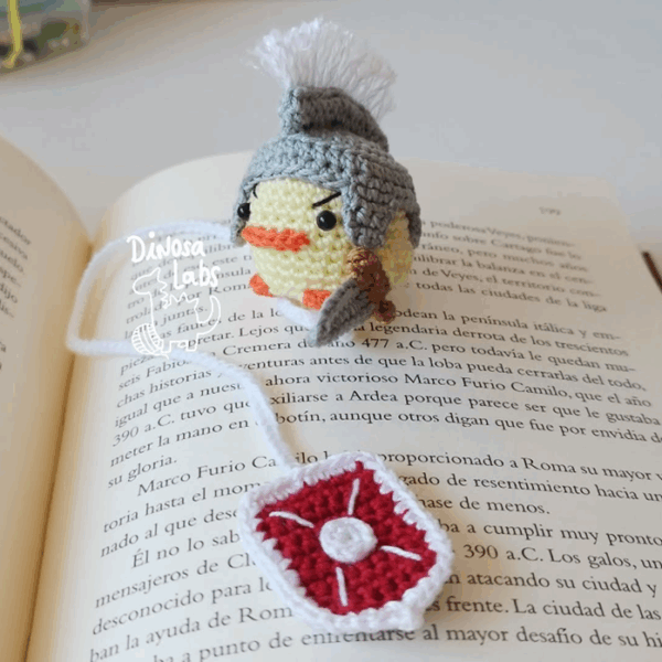 Amigurumi de un pollito vestido como un guerrero con un escudo con un hilo que sirve como marcapáginas. Se muestra sobre un libro