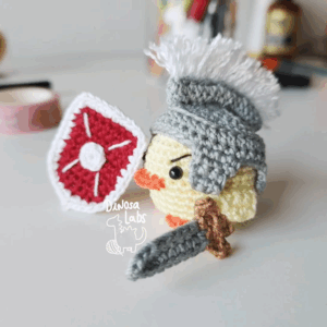 Amigurumi de un pollito vestido como un guerrero con un escudo con un hilo que sirve como marcapáginas
