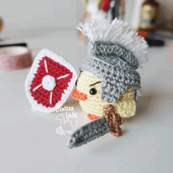 Amigurumi de un pollito vestido como un guerrero con un escudo con un hilo que sirve como marcapáginas