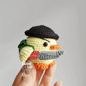 Vista lateral de un pollo tejido con crochet, tiene un cuchillo en un ala, y un gorro negro